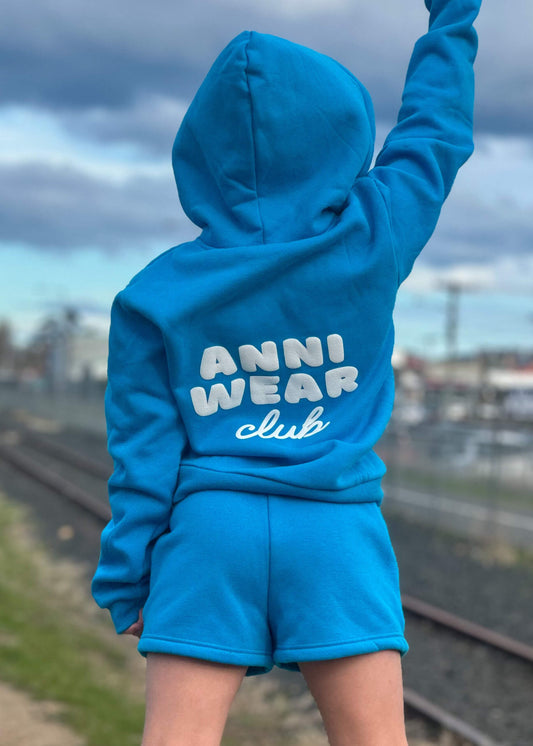 Person wearing a blue hoodie and shorts with 'Anni Wear Club' text, standing outdoors.