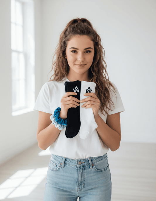 Woman holding a black and white sock with a logo, two scrunchies on wrist, wearing a white t-shirt and blue jeans, in a bright room.