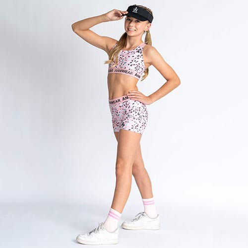 A female model wearing pink fall shorts with a pattern, paired with a pink and black patterned sports top and white sneakers. The model is posing with one hand on her hip.