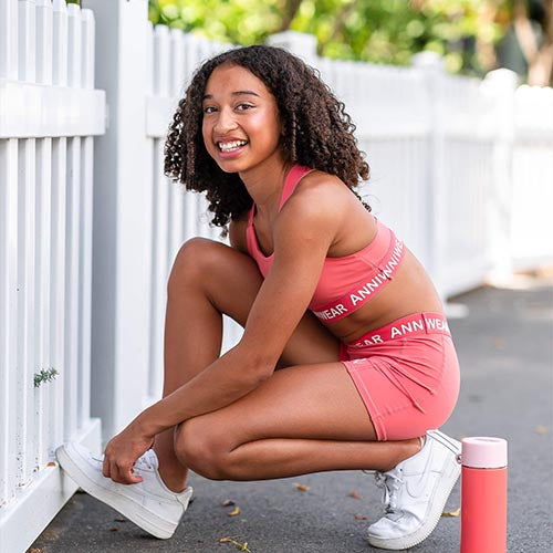 Woman in pink athletic wear posing outdoors with a white picket fence and greenery in the background