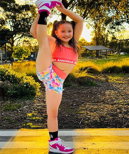 Young girl in colorful athletic wear performing a high leg stretch outdoors.