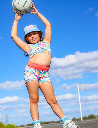 girl wearing a colorful crop top and shorts holding a volleyball outdoors with a cloudy sky background.