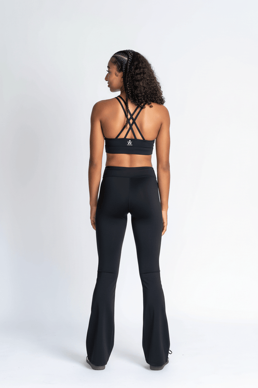 A girl is shown from the back wearing a black strappy sports bra and matching black high-waisted flared leggings against a plain white studio background.