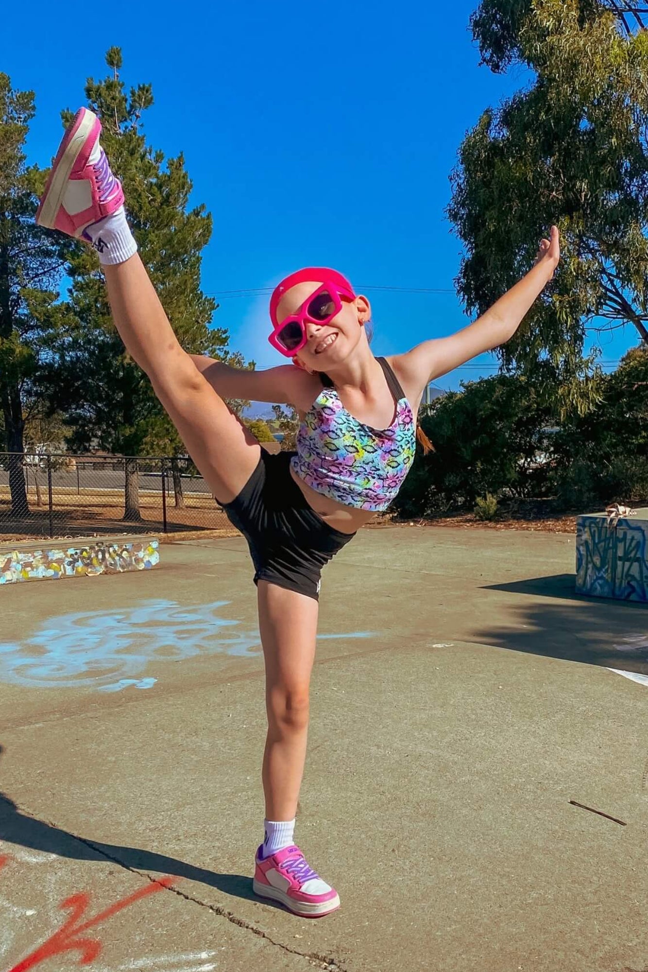 Person in colorful outfit and pink sunglasses posing outdoors with trees and clear sky in the background