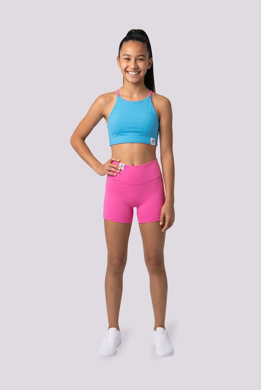 A young girl stands against a light gray studio background wearing a bright blue athletic crop top and high-waisted hot pink athletic shorts, paired with white sneakers.