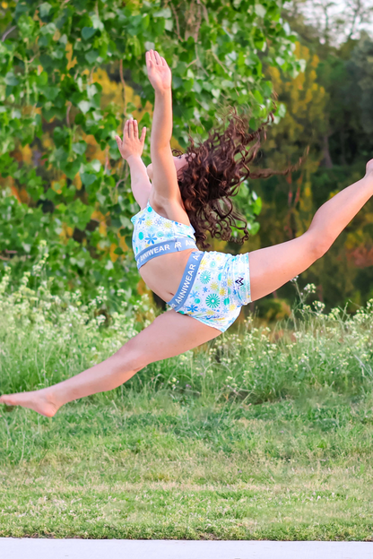 Person in a blue outfit performing a split leap in a grassy outdoor setting with trees and water in the background.