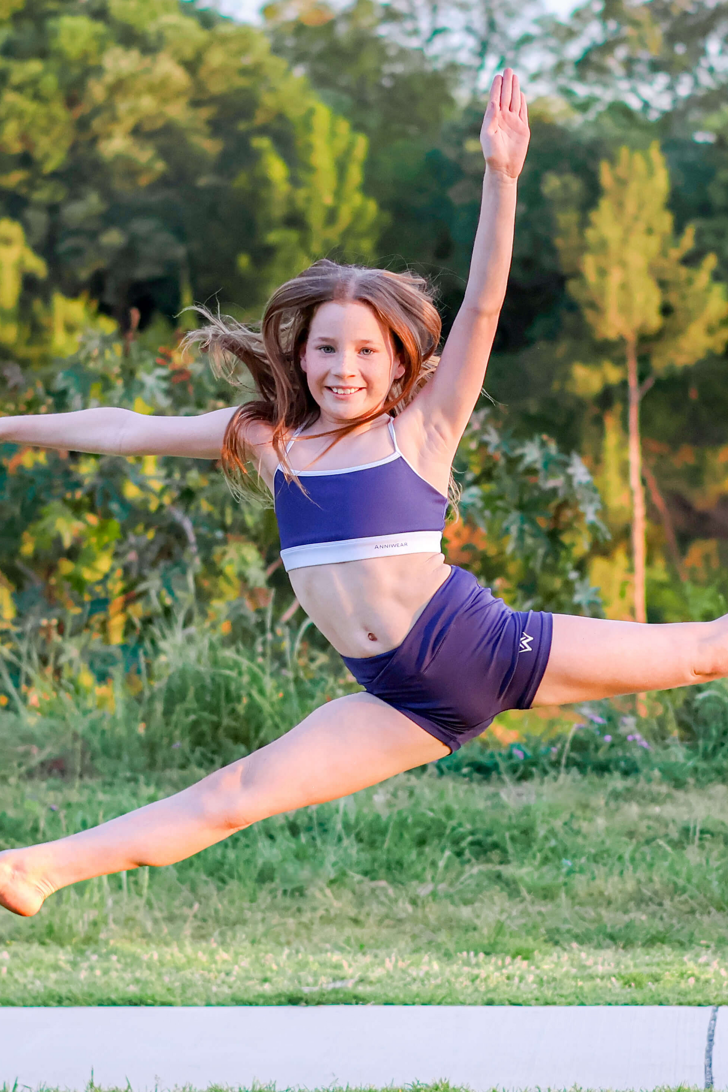Young girl in a blue and white outfit performing a split leap in a park.
