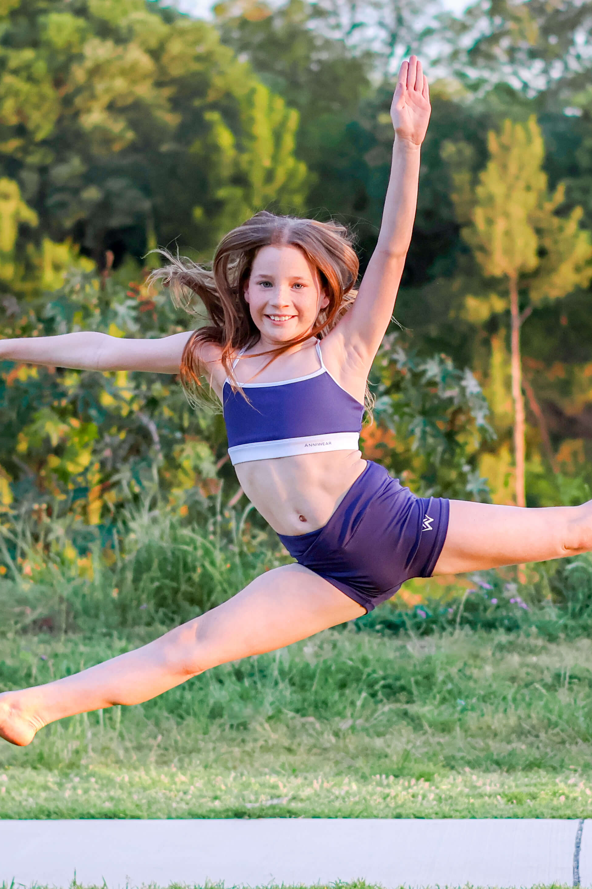 Young girl in a blue and white outfit performing a split leap in a park.