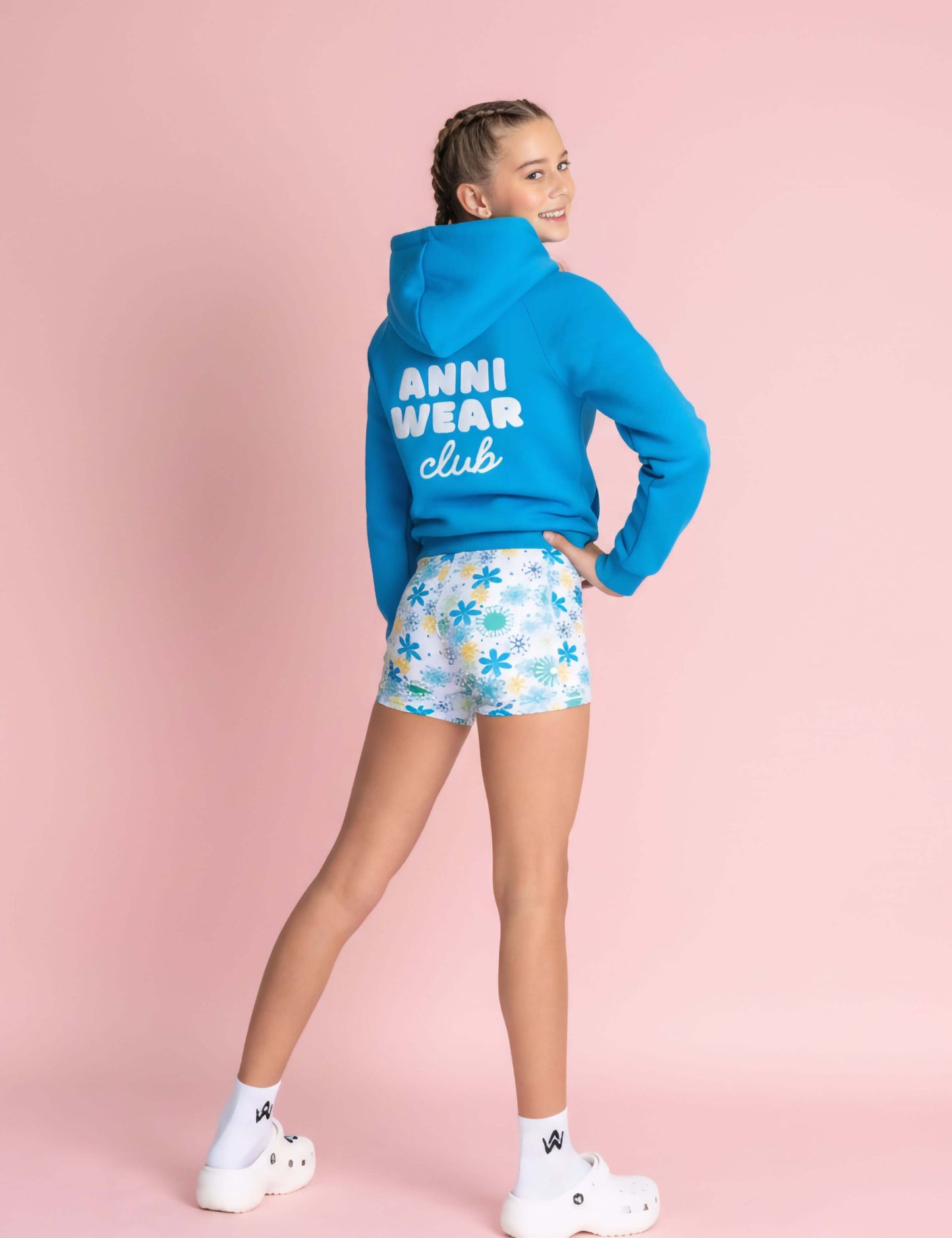 Person wearing a blue hoodie with 'ANNI WEAR club' text and patterned shorts on a pink background