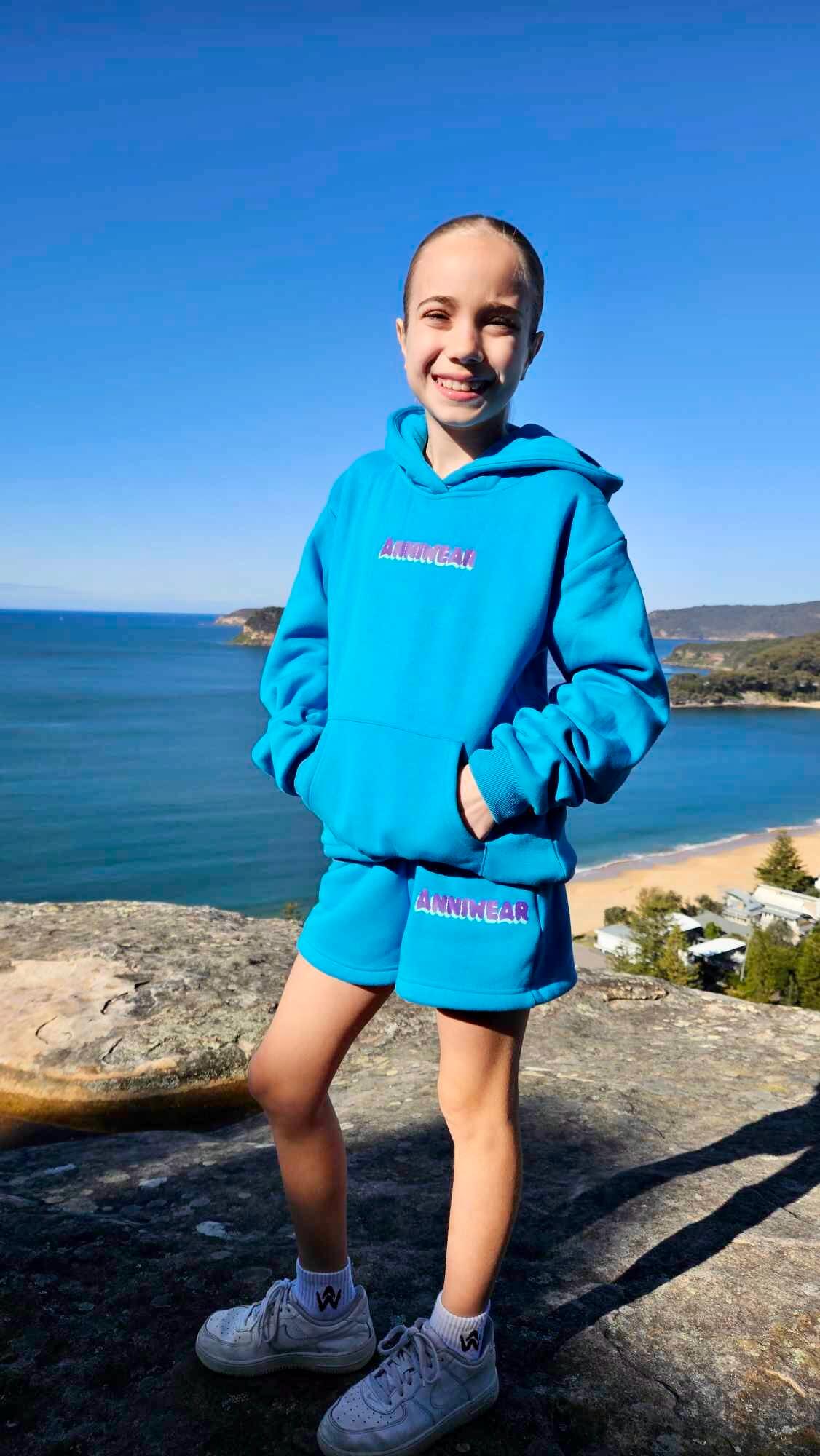Person wearing a blue hoodie and shorts set with text, standing on a rocky outcrop with ocean view.