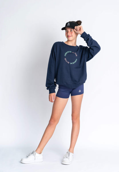 A female model wearing a navy blue French terry jumper with a relaxed fit and dropped shoulders, paired with navy blue shorts. She is posing with one hand on her hip and the other hand raised to her head.