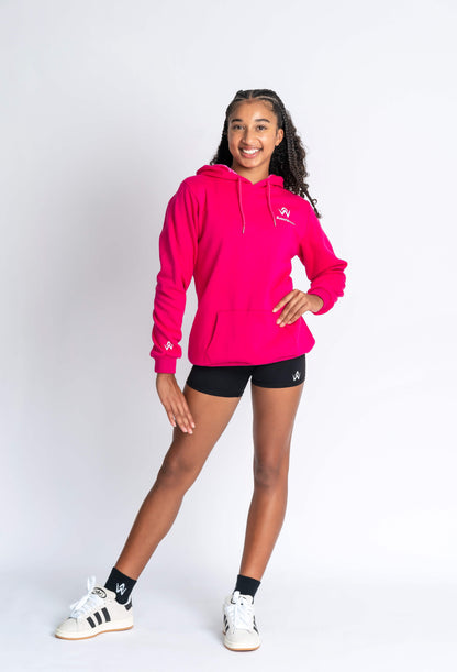 A child model wearing a hot pink hoodie with a logo on the chest and black shorts, standing against a white background.