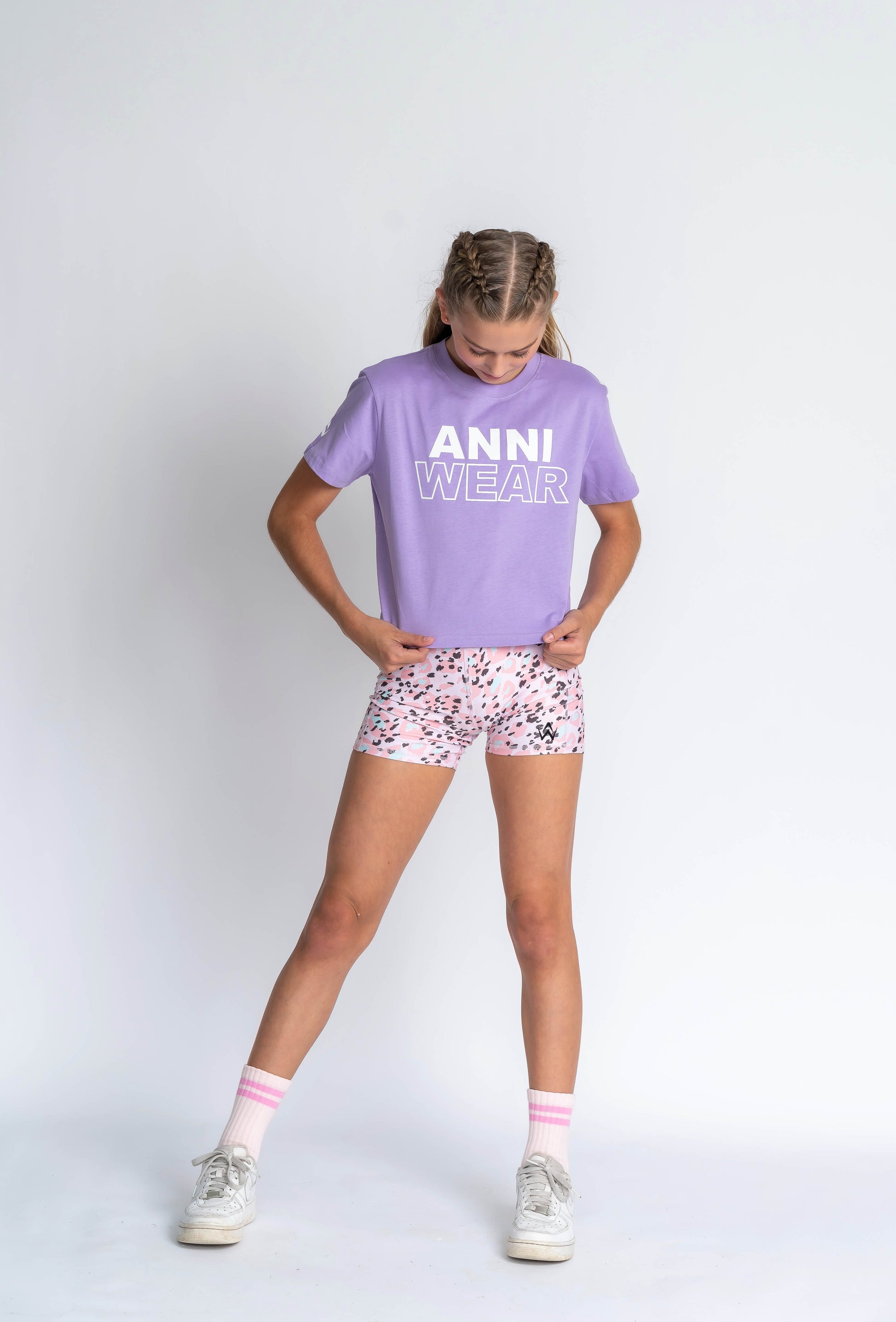 A person wearing a lavender crop style tee with a white chest signature print, and matching pink shorts, standing against a plain background.