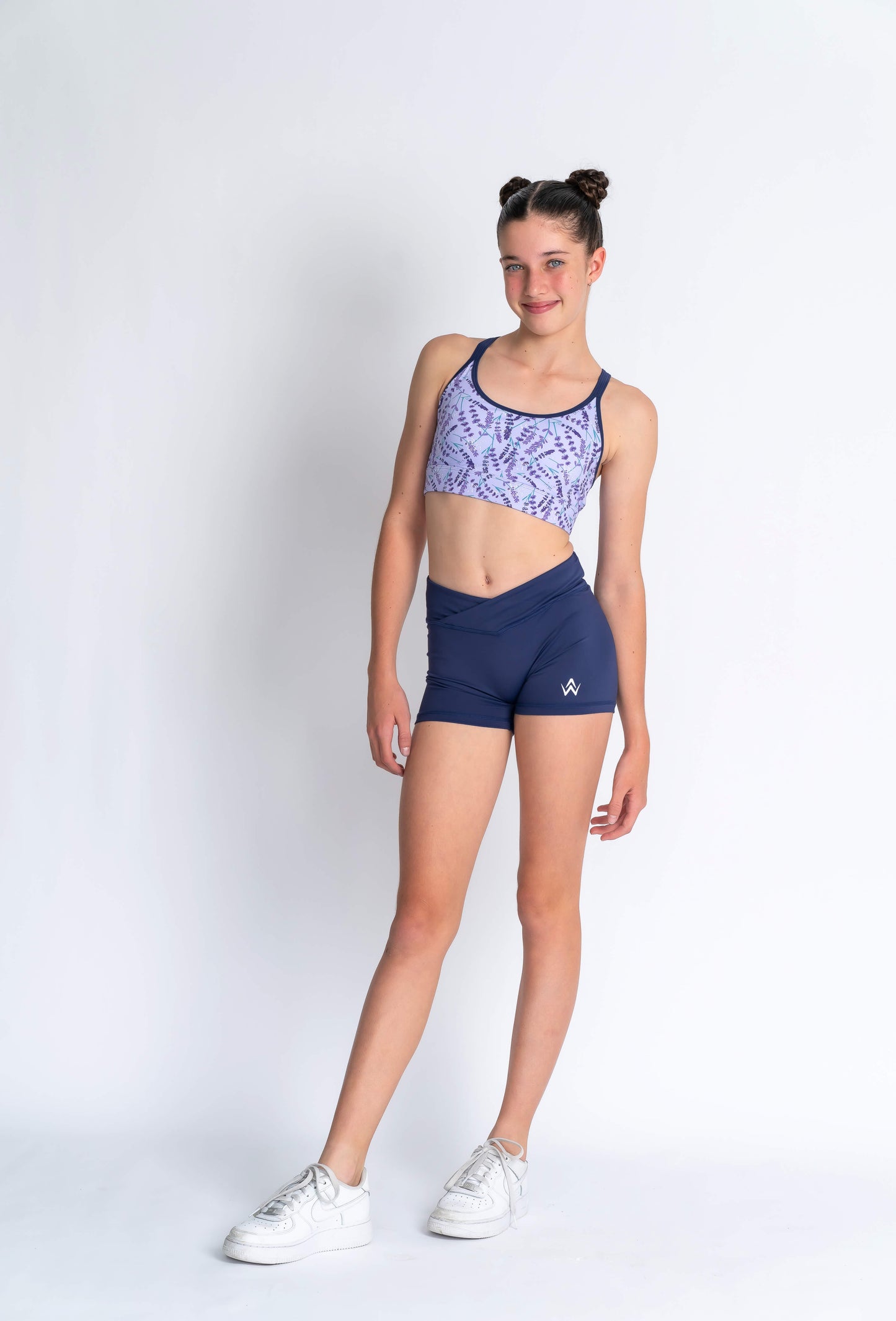 A female model wearing a lavender haze crop top with a patterned design, paired with navy blue shorts, white sneakers, and has her hair styled in a bun.