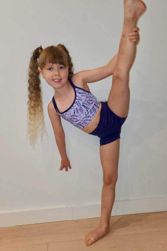 Young girl in a gymnastics pose with one leg extended on a wooden floor against a white wall.