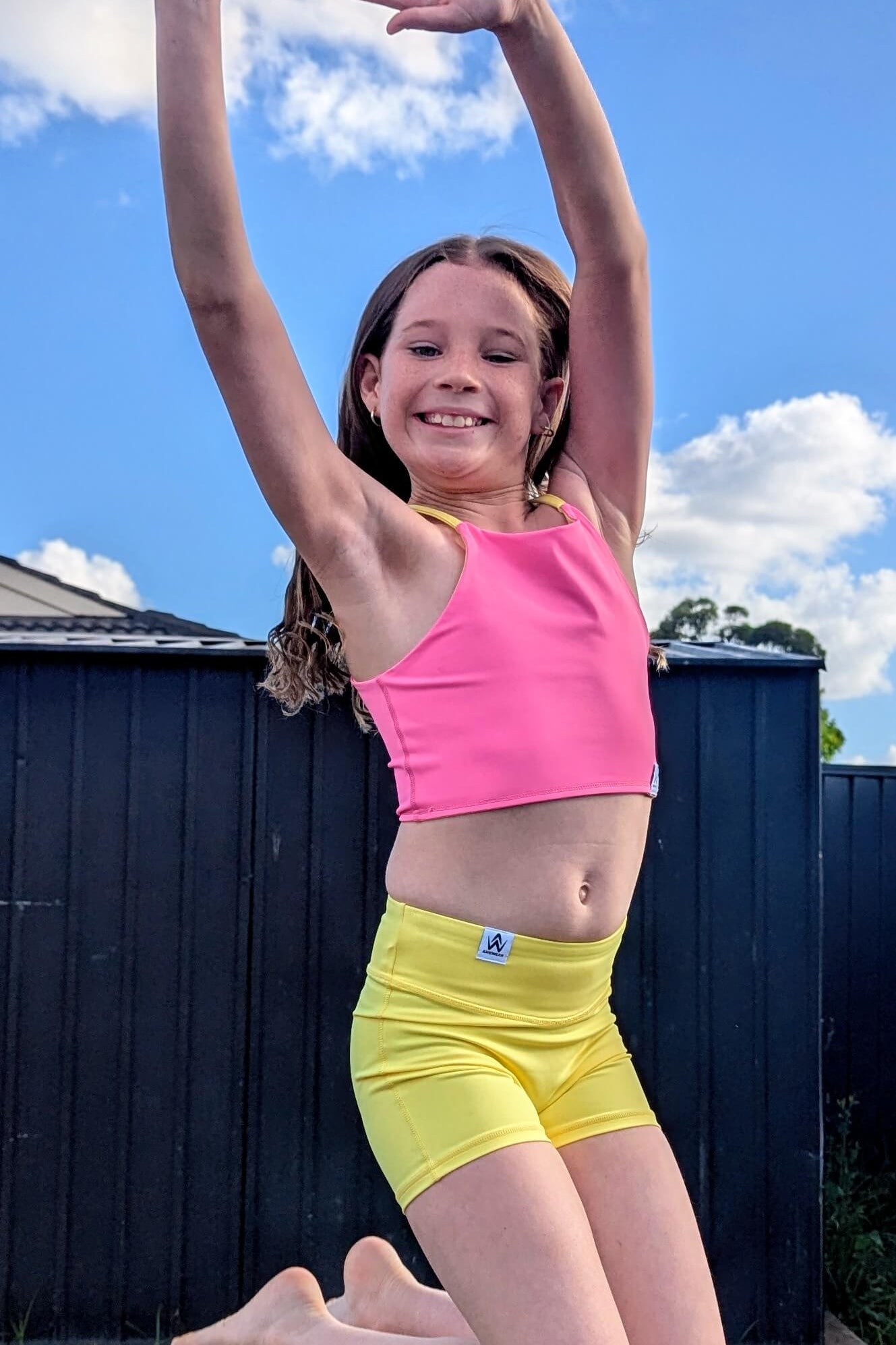 Young girl in a pink sports bra and yellow shorts jumping outdoors with a blue sky and white clouds in the background.