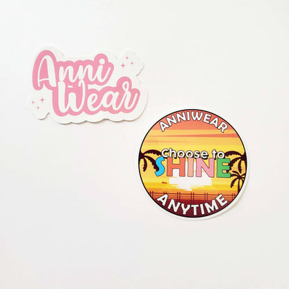 Sticker with Pink 'Anni Wear' text on a white background. and Circular sticker with sunset design and text 'Choose to Shine Anytime' on a white background
