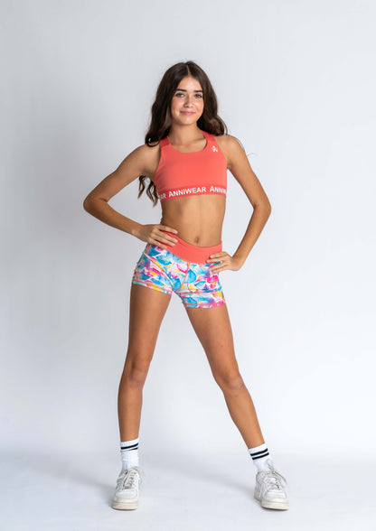 A female model wearing a coral sports crop top and multicolored tropical sports shorts.