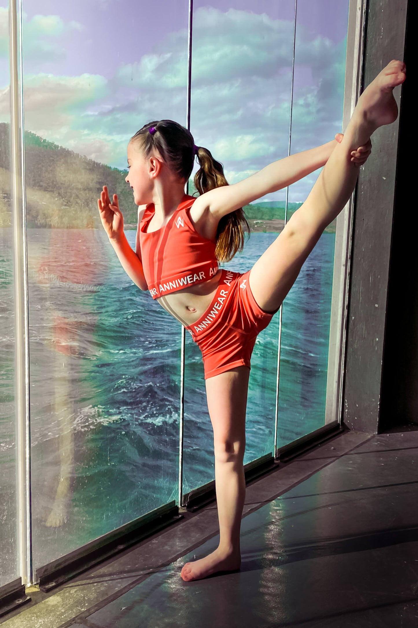 Young girl in red athletic outfit stretching by a large window with a scenic view.