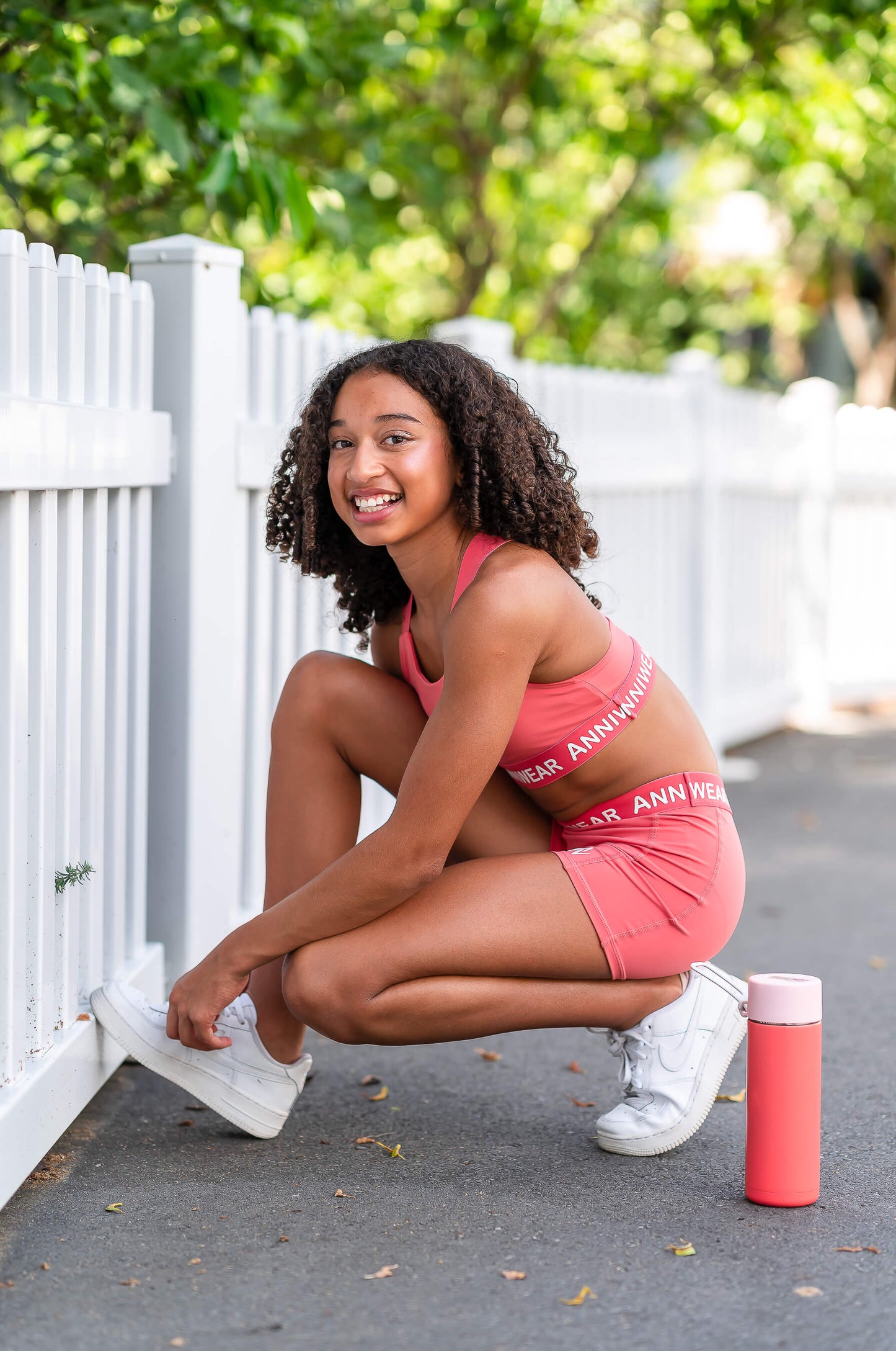 Woman in pink athletic wear posing outdoors with a white picket fence and greenery in the background