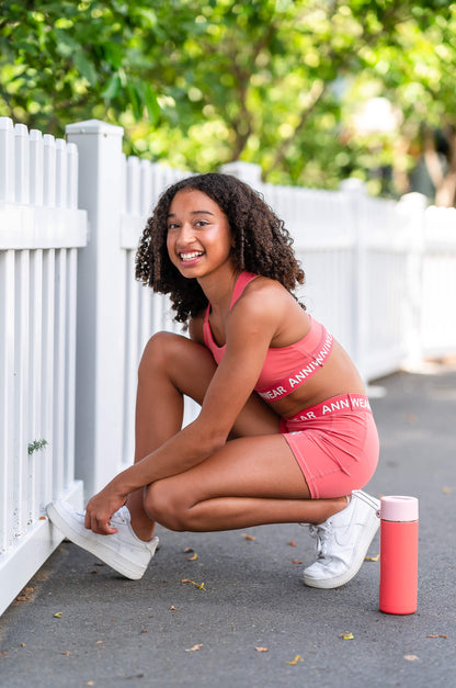 Woman in pink athletic wear posing outdoors with a white picket fence and greenery in the background