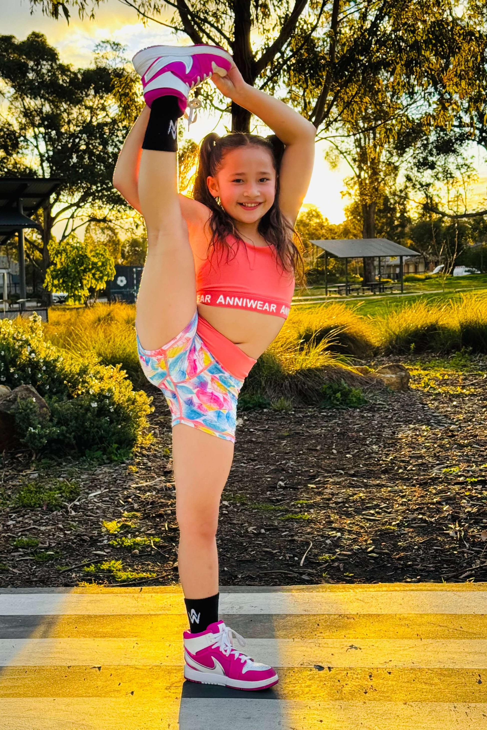 Young girl in colorful athletic wear performing a high leg stretch outdoors.