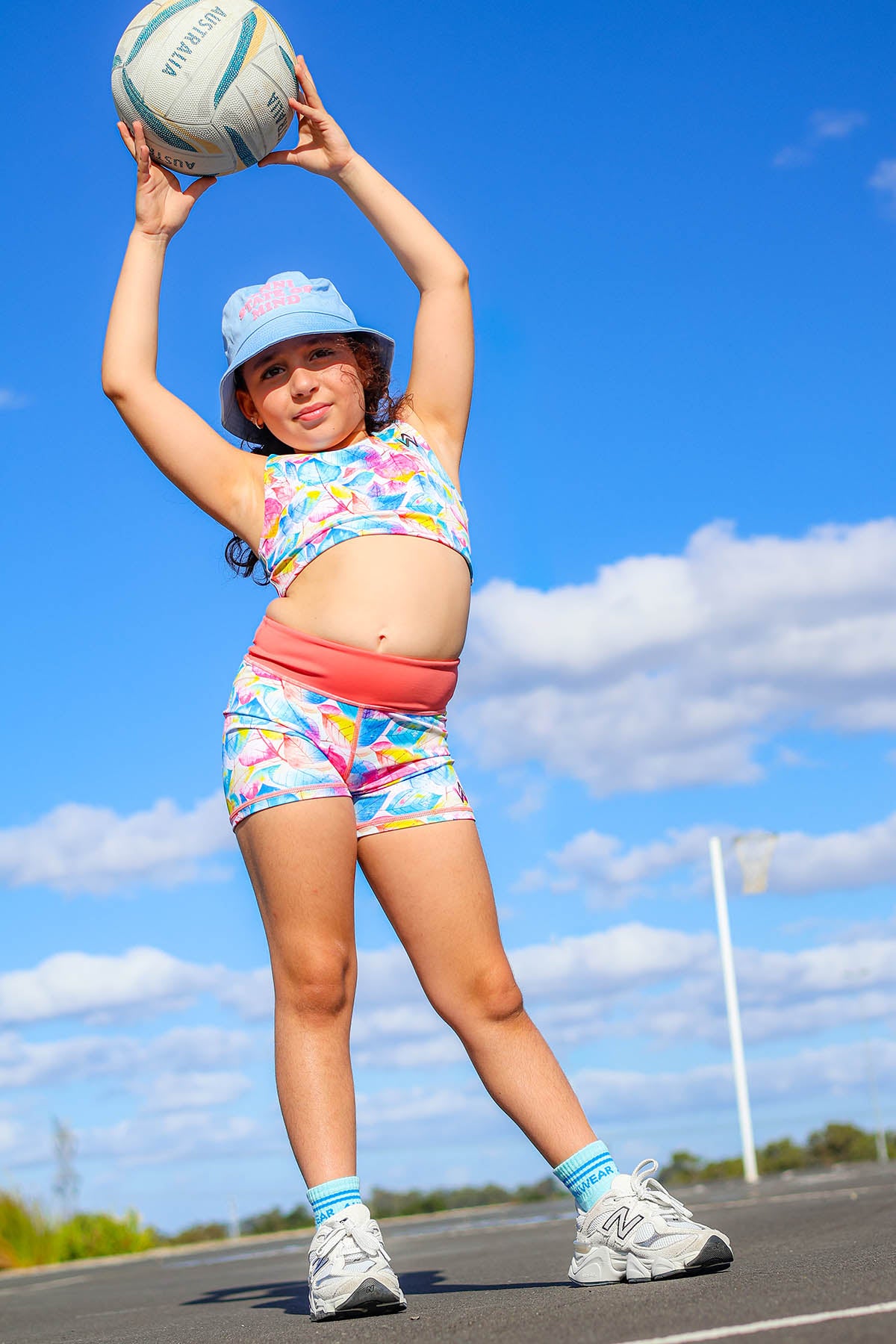 girl wearing a colorful crop top and shorts holding a volleyball outdoors with a cloudy sky background.