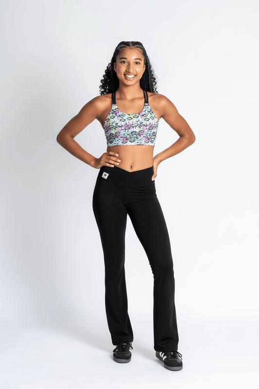 A young girl stands in a studio wearing a floral print sports bra and black flared leggings with black sneakers, photographed against a light gray background.